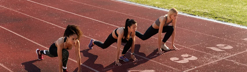 Three female athletes preparing for a sprint race at the starting line of an outdoor athletics track.